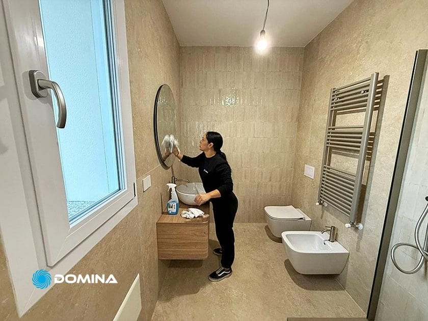 Una persona vestita di nero pulisce lo specchio rotondo del bagno con un panno. I prodotti per la pulizia sono sul lavandino. Il bagno di Rho è caratterizzato da piastrelle beige, un radiatore per asciugamani, un bidet e un WC-Casa Pronta dopo Pulizie Fine Cantiere.