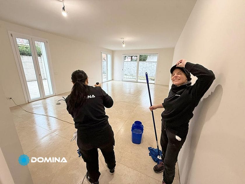 Due donne stanno pulendo una stanza vuota e luminosa a Rho, con grandi finestre e pavimento piastrellato. Una donna sorride e si stiracchia tenendo in mano uno spazzolone; l'altra, impegnata con le pulizie a fine cantiere, dà le spalle all'obiettivo.