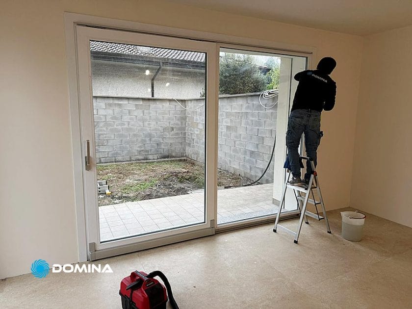 Una persona è in piedi su una scaletta vicino a una grande porta scorrevole di vetro e sta eseguendo Pulizie Fine Cantiere. All'esterno si intravede un piccolo cortile e un muro di cemento, mentre all'interno una cassetta degli attrezzi rossa e un secchio bianco sono appoggiati sul pavimento, segno di Casa Pronta a Rho.