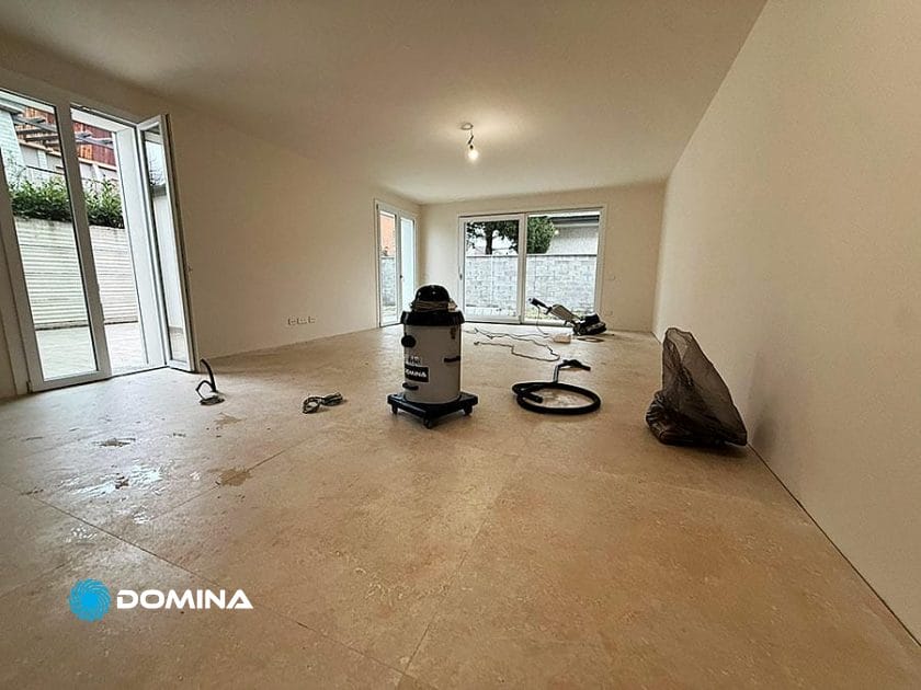 Una stanza per lo più vuota con pavimenti in piastrelle, grandi finestre e un'illuminazione minima. Attrezzature per la pulizia, come aspirapolvere e attrezzi, sono sparsi sul pavimento, segno di Pulizie Fine Cantiere, mentre il processo di Casa Pronta prepara lo spazio per un nuovo inizio.