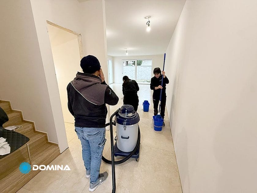 Alla Casa Pronta di Rho, tre persone eseguono Pulizie Fine Cantiere in una stanza vuota e appena ristrutturata: uno passa l'aspirapolvere sul pavimento, mentre altri due passano lo straccio e raccolgono le provviste vicino alla porta, mettendo in risalto pavimenti e pareti chiare e immacolate.