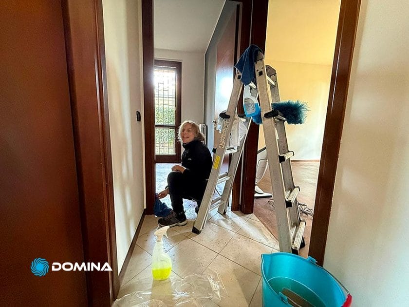 Un bambino sorridente è seduto a terra in un corridoio accanto a una scala a pioli argentata, a materiale per le pulizie e a un secchio blu, forse aiutando con Pulizie Straordinarie Appartamento a Lainate o altri compiti di manutenzione domestica.
