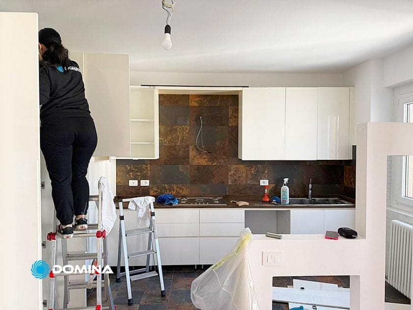 Una persona è in piedi su una scala a pioli e sta entrando in un mobile superiore di una cucina moderna. La scena suggerisce Pulizie Straordinarie Appartamento a Lainate, con materiale di pulizia sui banconi e alcuni pannelli rimossi per una pulizia approfondita.