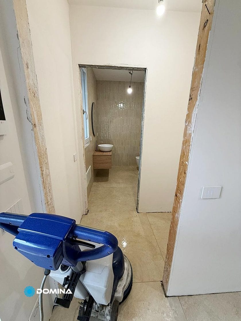 Un bagno parzialmente ristrutturato a Rho presenta pavimenti piastrellati beige, una parete bianca, un piccolo lavandino con mobiletto in legno e una macchina lavapavimenti Pulizie Fine Cantiere. I telai delle porte a vista e una lampadina spoglia contribuiscono a creare un'atmosfera da Casa Pronta.