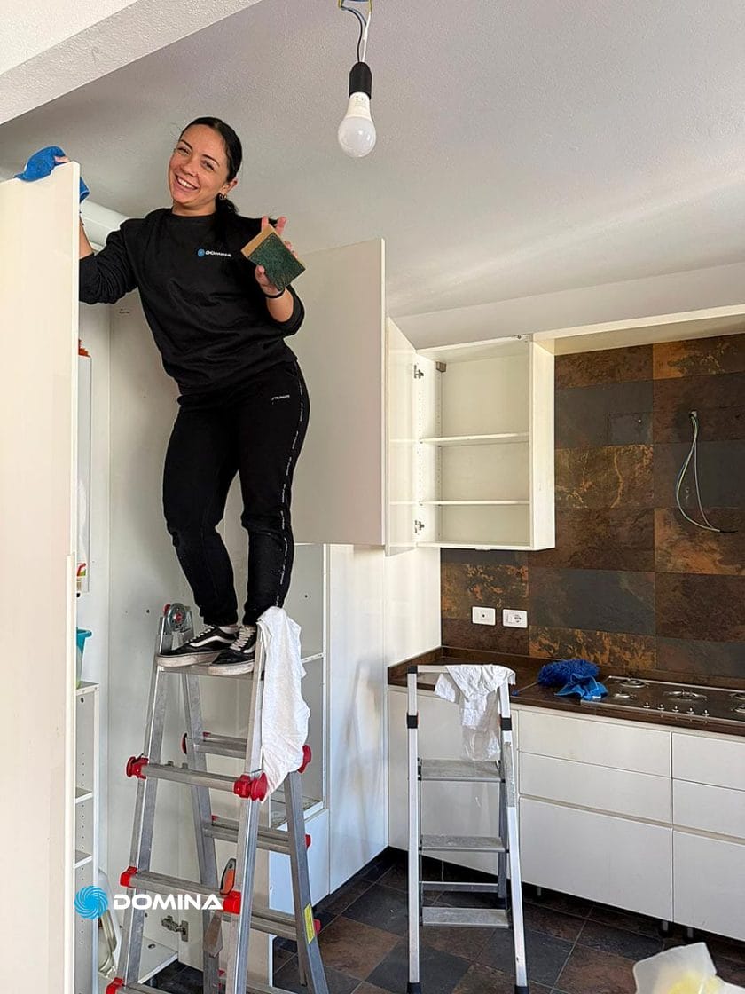 Una donna è in piedi su una scala e sorride mentre pulisce un mobile bianco della cucina, mostrando Pulizie Straordinarie Appartamento a Lainate. La luminosa cucina presenta armadietti aperti, banconi bianchi, pareti piastrellate, un'altra scala e panni per la pulizia a vista.