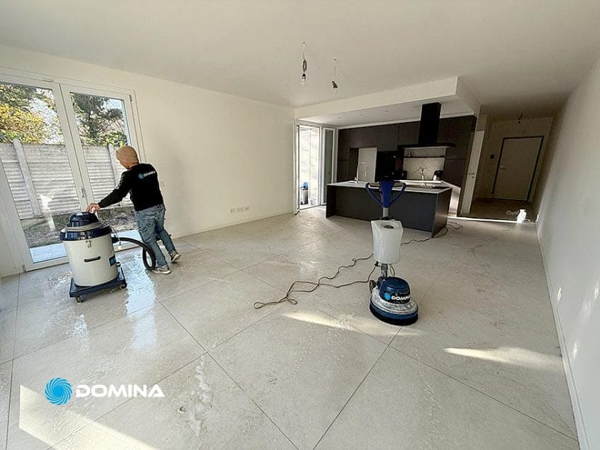 Una persona utilizza un aspirapolvere per pulire l'ampio pavimento piastrellato di una luminosa e moderna cucina e zona giorno a Origgio, eseguendo pulizie post ristrutturazione con attrezzature di pulizia visibili nella stanza.