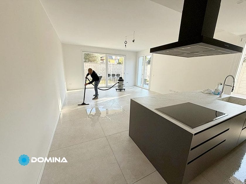 Una persona utilizza una macchina lavapavimenti per pulire il pavimento in piastrelle di una moderna cucina e zona giorno non arredata a Origgio dopo la pulizie post ristrutturazione, con pareti bianche e grandi finestre.