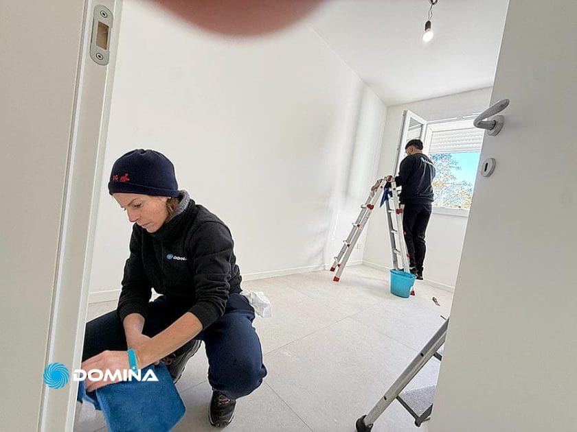Due persone stanno pulendo una stanza bianca e luminosa dopo Pulizie Post Ristrutturazione. Una è inginocchiata vicino alla porta e pulisce il pavimento, mentre l'altra è in piedi vicino a una finestra aperta su una scala a pioli con un secchio blu. Una mano copre parzialmente l'obiettivo della telecamera.