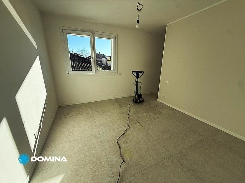 Una stanza vuota a Origgio con pareti e pavimento chiari, la luce del sole che filtra dalla finestra e una macchina lavapavimenti inserita, ideale per le pulizie post ristrutturazione dopo il lavoro in cantiere.