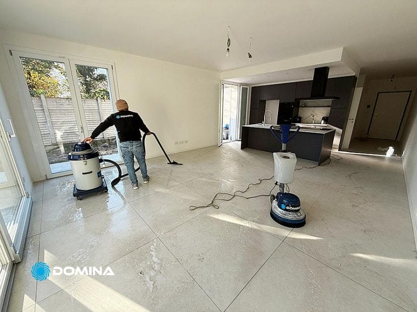 Un operaio sta eseguendo pulizie post ristrutturazione su un pavimento piastrellato spazioso e moderno in una stanza luminosa con grandi finestre a Origgio. Sono visibili le attrezzature per la pulizia e un macchinario, con una zona cucina aperta sullo sfondo.