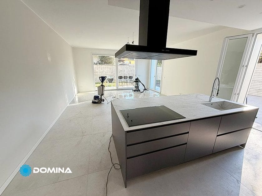 Una moderna cucina non arredata a Origgio con isola in marmo, piano cottura a induzione e lavello. Grandi porte scorrevoli in vetro illuminano il pavimento piastrellato, dove sullo sfondo sono visibili le macchine per la pulizia Pulizie Post Ristrutturazione.