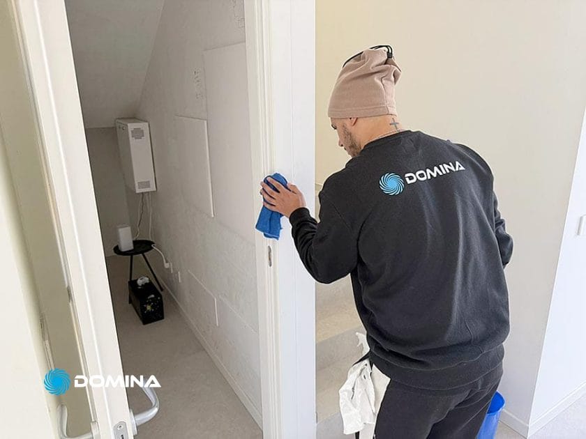 Una persona che indossa un berretto e una felpa "DOMINA" pulisce con un panno blu il telaio di una porta bianca in una stanza luminosa e ordinata dopo le pulizie post ristrutturazione in un cantiere di Origgio.
