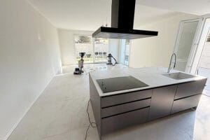 Una cucina moderna e minimale a Origgio con isola in marmo, piano cottura a incasso e cappa aspirante. Le grandi piastrelle del pavimento e le attrezzature per la pulizia mostrano la cura di Pulizie Post Ristrutturazione, mentre le porte di vetro si aprono su un invitante spazio esterno.