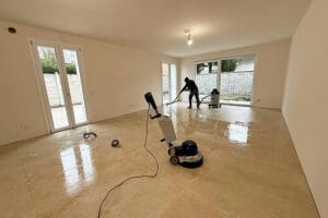 Due persone stanno pulendo il pavimento in piastrelle di una stanza vuota e non arredata a Rho, piena di luce naturale. Utilizzando macchine lavapavimenti, eseguono Pulizie Fine Cantiere per garantire che lo spazio sia immacolato e pronto per Casa Pronta.