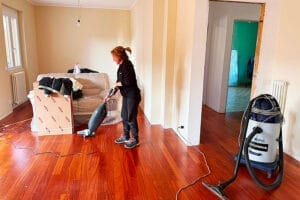 Una donna passa l'aspirapolvere sul parquet in una stanza per lo più vuota, preparandosi per Pulizie Straordinarie Appartamento a Lainate. Il divano è ricoperto di plastica, le scatole per il trasloco sono vicine e un grande aspirapolvere è collegato alle pareti nude.