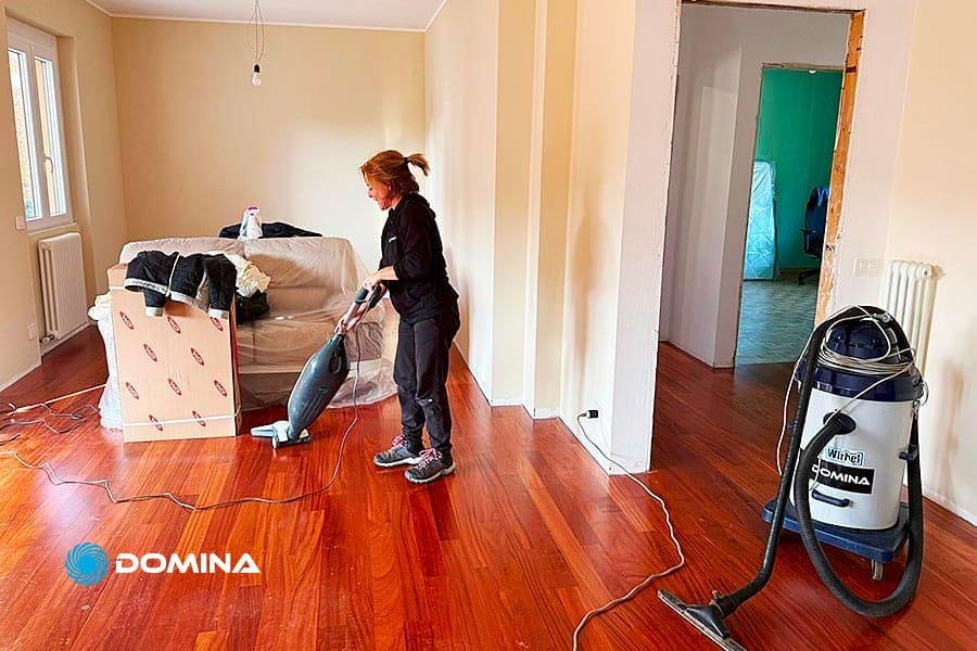 Una donna passa l'aspirapolvere sul parquet in una stanza per lo più vuota, preparandosi per Pulizie Straordinarie Appartamento a Lainate. Il divano è ricoperto di plastica, le scatole per il trasloco sono vicine e un grande aspirapolvere è collegato alle pareti nude.